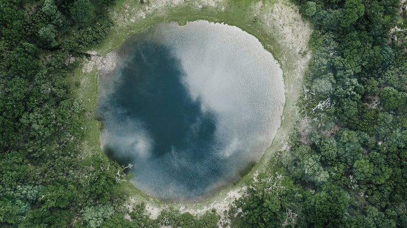 En rund sjö fotograferad i helikopterperspektiv, med omgärdande skog.